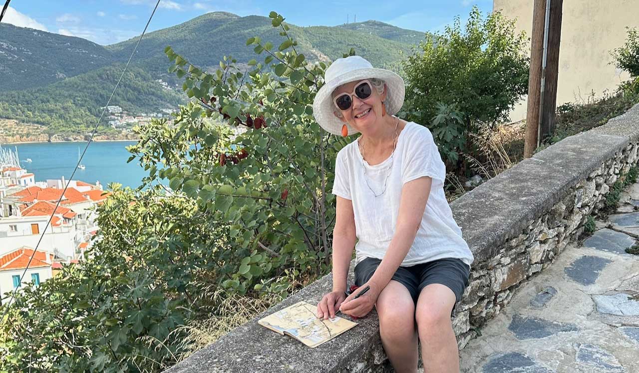 Debbie Mackinnon wearing a sun hat sitting on a wall, sketching in Greece. Sea and hills in the background.
