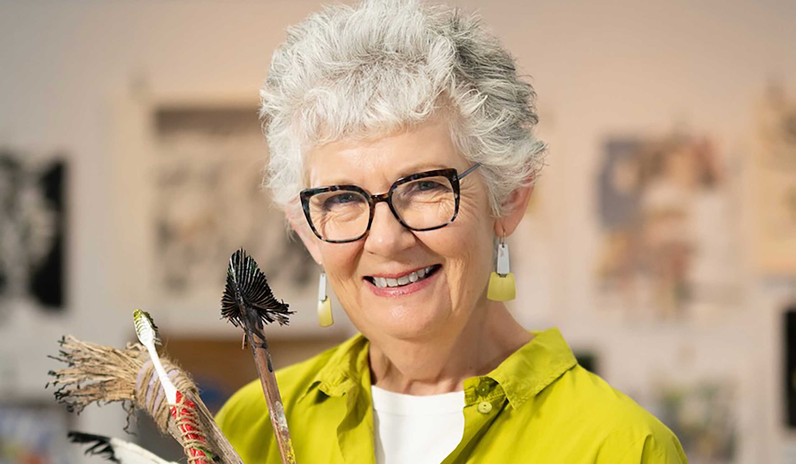 Debbie Mackinnon wearing a yellow blouse and smiling into the camera whilst holding paint brushes