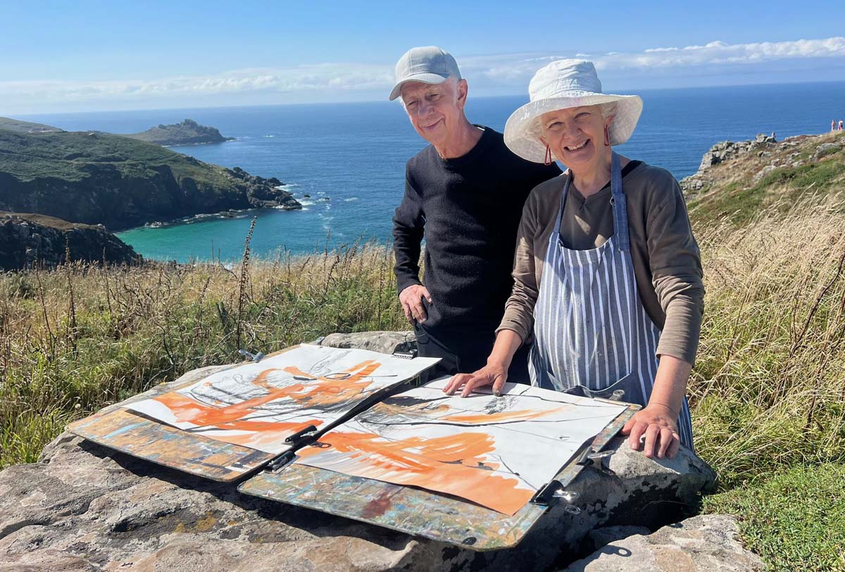 Two people stand outdoors by a rocky coastline with hills and the sea in the background. They are posing behind two sketchpads on a stone ledge, displaying orange and pencil drawings. Both are smiling and wearing hats.