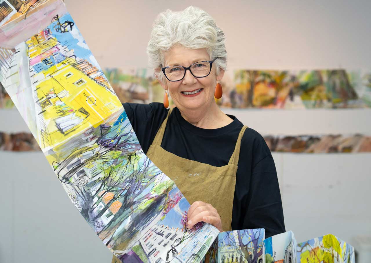 A woman with short grey hair, glasses, and orange earrings smiles whilst holding a long, colourful illustrated accordion sketch book. She is wearing a brown apron over a black shirt and standing in a brightly lit art studio.