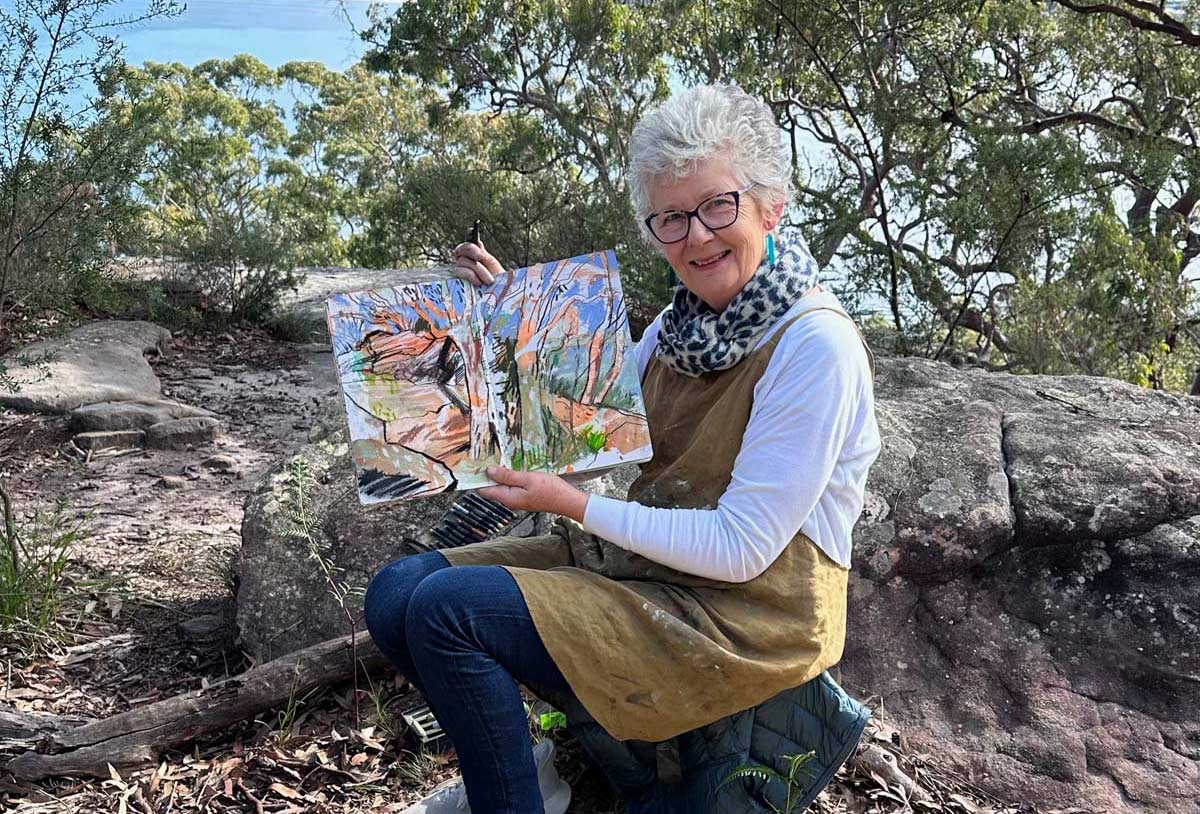 A person with short grey hair, glasses, and an apron sits outdoors on a rock, holding an open sketchbook with a colourful landscape painting, surrounded by trees and foliage.
